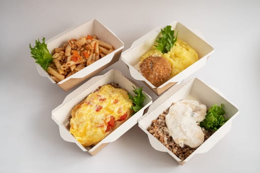 Four takeout meals served in biodegradable containers, featuring pasta, casserole, mashed potatoes with meatball, and buckwheat with chicken, garnished with greens.