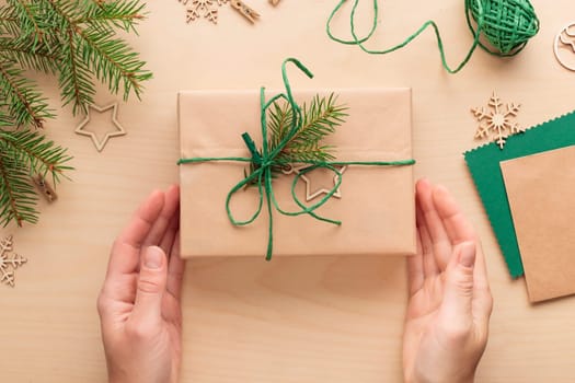 Female hands holding Merry Christmas eco-friendly natural packaging gift box on wooden decorated festive background, woman prepare a surprise for the new year, winter handmade holiday green present concept, flat lay, top view