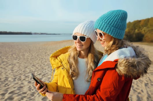 Happy modern young two women friends with smartphone together on beach, stylish smiling girlfriends using phone on sea coast in hat, jacket outdoors on a sunny warm day