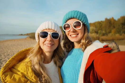 Happy joyful young two women friends taking selfie with smartphone together on sunny beach, stylish smiling girlfriends having fun on sea coast in glasses, jacket, hat outdoors