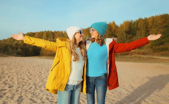 Happy joyful young two women friends together on sunny beach, stylish smiling girlfriends standing on sea coast in glasses, colorful red yellow coat jacket and hat outdoors