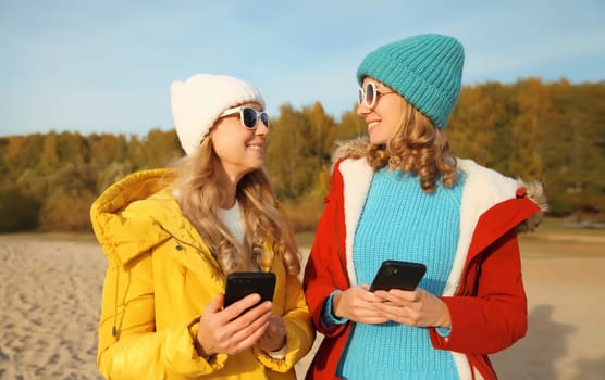Happy modern young two women friends with smartphone together on beach, stylish smiling girlfriends using phone on sea coast in hat, jacket outdoors on a sunny warm day