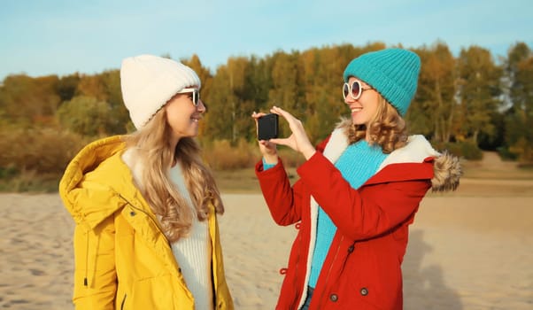 Happy joyful young two women friends taking selfie with smartphone together on sunny beach, stylish smiling girlfriends having fun on sea coast in glasses, jacket, hat outdoors