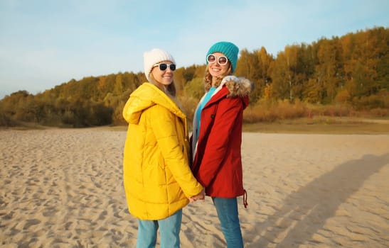 Happy young two women friends together on sunny beach, stylish smiling girlfriends standing on sea coast in glasses, colorful red yellow coat jacket and hat outdoors