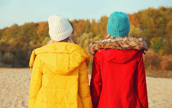 Back view of two young modern women friends standing together on sunny beach, stylish girlfriends on sea coast in colorful red yellow coat jacket and hat outdoors