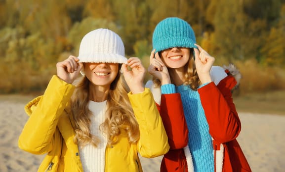 Portrait of happy two women friends having fun pulling knitted winter colorful hat over her face covering her eyes, funny girlfriends together outdoors in park