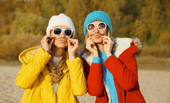 Portrait of happy two women friends having fun showing mustache in winter colorful hat, glasses, funny girlfriends together outdoors in park