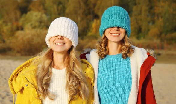 Portrait of happy two women friends having fun pulling knitted winter colorful hat over her face covering her eyes, funny girlfriends together outdoors in park