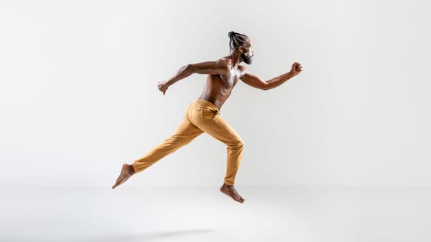 Determined shirtless gay model jumping over gray background. Full body side view of strong young man is in midair at plain studio.