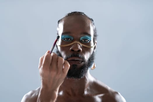 Confident African American gay model applying eye makeup over gray background. Portrait of serious homosexual man with chain on nose in studio.