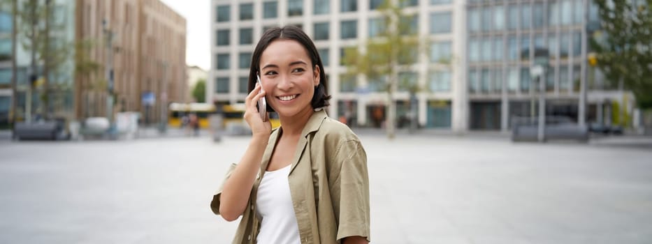 Modern young asian girl talks on mobile phone, uses telephone on city street. Woman smiling while calling someone on smartphone.