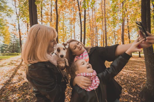Mother, grandmother and little granddaughter with jack russell terrier dog taking selfie by smartphone outdoors in autumn nature. Family, pets and generation concept.