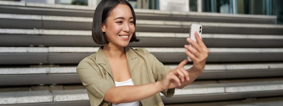 Portrait of asian beautiful girl takes selfie on smartphone. Young korean woman sits on stairs outdoors and makes photos on mobile phone.