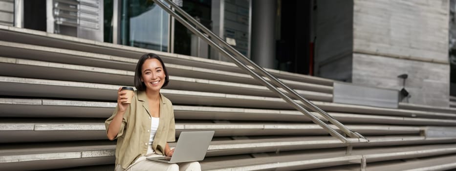 Vertical shot of smiling girl student, asian woman sits on stairs of university campus and drinks coffee, does her homework on laptop.