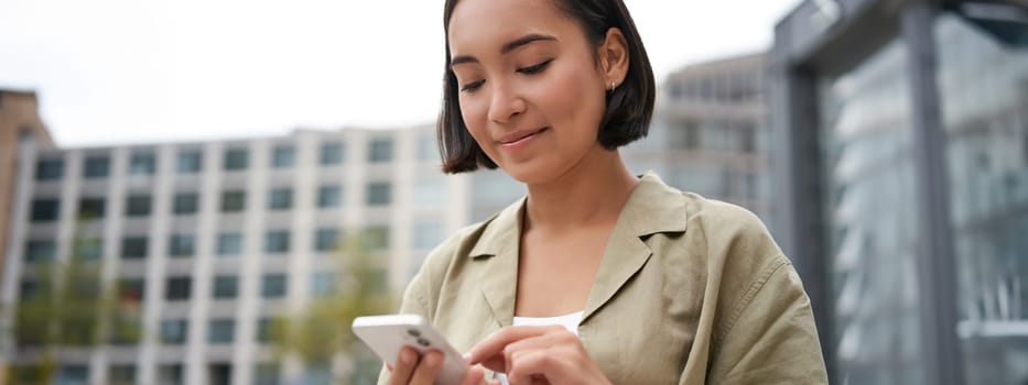 Mobile technology. Smiling asian woman using smartphone app, looking at her telephone on street, checking map, calling or texting someone.