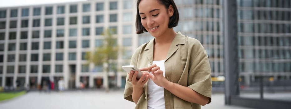 Mobile technology. Smiling asian woman using smartphone app, looking at her telephone on street, checking map, calling or texting someone.