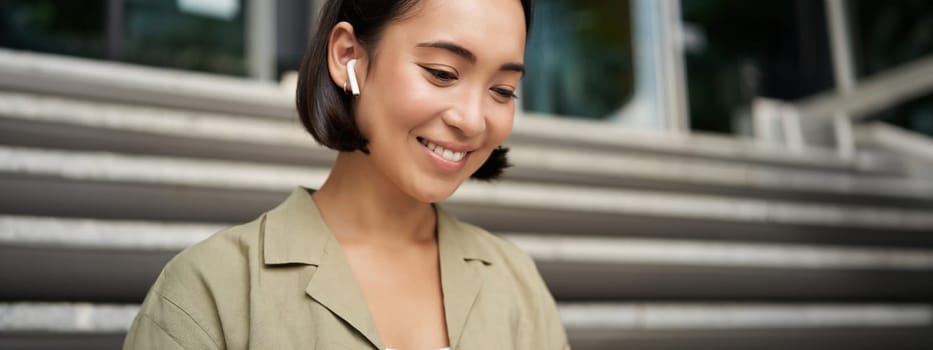 Portrait of smiling asian girl listens music, podast in wireless earphones, using headphones outdoors, sitting on street.