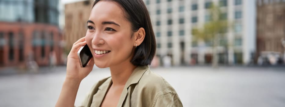 Modern young asian girl talks on mobile phone, uses telephone on city street. Woman smiling while calling someone on smartphone.