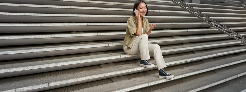 Smiling asian girl sits on stairs of building and talks on mobile phone, relaxing during telephone conversation.