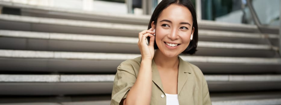 Close up portrait, street photo of asian woman listening music in wireless headphones, smiling, sitting on stairs.