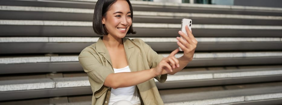 Portrait of asian beautiful girl takes selfie on smartphone. Young korean woman sits on stairs outdoors and makes photos on mobile phone.