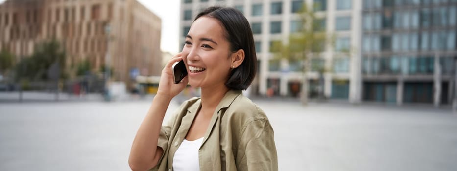 Modern young asian girl talks on mobile phone, uses telephone on city street. Woman smiling while calling someone on smartphone.