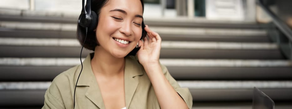Happy asian girl listens to music in headphones, smiles, sits outdoors on stairs.