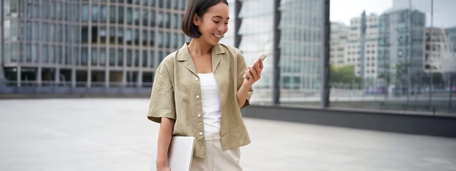 Young asian woman with laptop, walking on street and texting message, smiling while looking at smartphone.