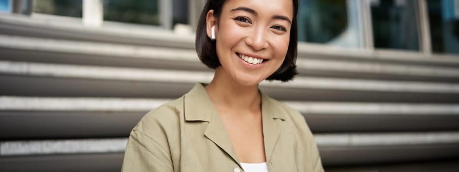 Portrait of smiling asian girl listens music, podast in wireless earphones, using headphones outdoors, sitting on street.