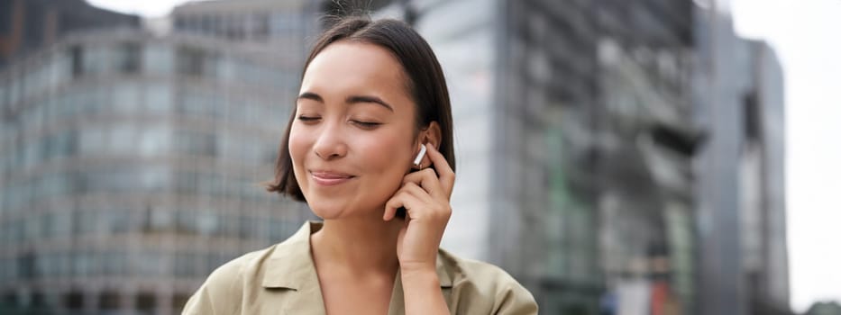 Vertical shot of smiling asian woman in wireless headphones, enjoys listening to music in earphones, holds mobile phone.