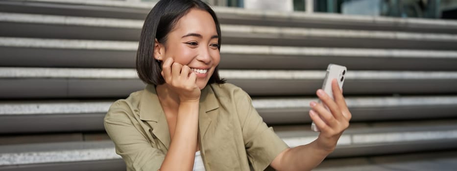 Portrait of happy smiling asian girl takes selfie on mobile phone. Young korean woman video chat on smartphone while sitting on stairs near building.