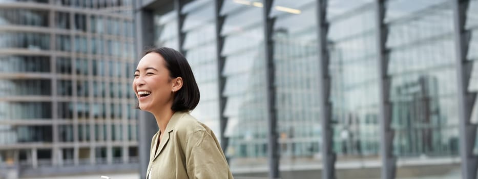 Vertical shot of beautiful asian woman walking on street, laughing and looking happy, enjoying the day.