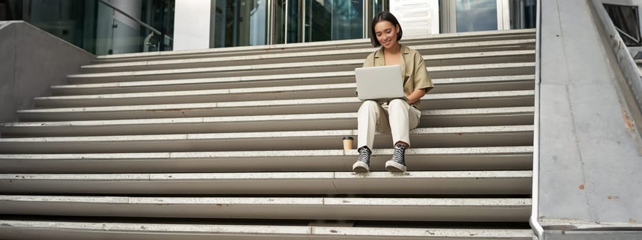 Asian girl student sits on stairs near campus, types on laptop, does her homework outdoors.