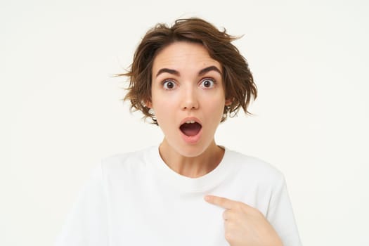 Close up portrait of girl with surprised face, points at herself, looks shocked, stands over white background.