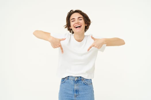 Portrait of cute, happy young woman pointing fingers down, showing advertisement, demonstrating banner on the bottom, standing against white background.