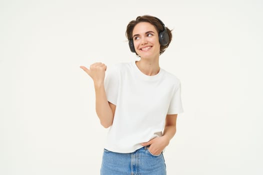 Image of young modern woman, wearing white t-shirt, pointing left, smiling and looking happy, showing advertisement, standing over white background.