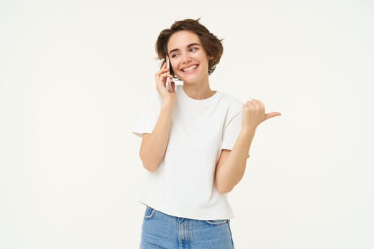 Portrait of young woman talking on mobile phone, using smartphone and pointing finger right, showing direction, banner advertisement, studio background.