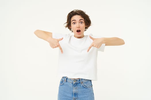 Portrait of amazed girl, pointing fingers down, showing something and looking surprised, standing over white studio background.