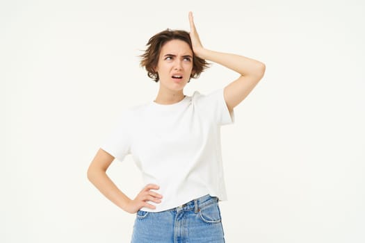 Portrait of woman looking troubled, slap her forehead, standing thoughtful and frowning, facing complicated problem, standing over white studio background.