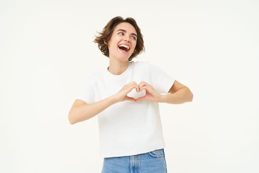 Portrait of carefree and happy woman, laughing, showing heart gesture near chest, expresses love and care, isolated over white background.