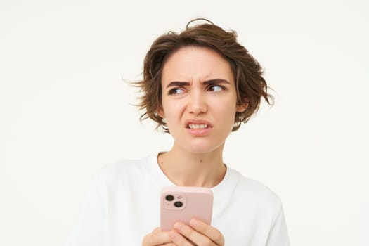 Portrait of puzzled brunette woman, standing with smartphone, thinking and frowning, looking reluctant, standing over white background.