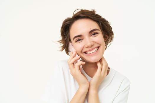 Image of smiling, brunette woman calling someone, talking on mobile phone, answer telephone call, standing over white background.