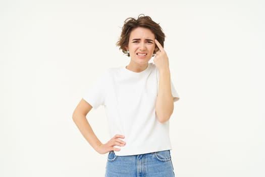 Portrait of woman reacting to smth riddiculous, pointing finger at forehead and frowning, complaining at someone stupid, standing over white background.