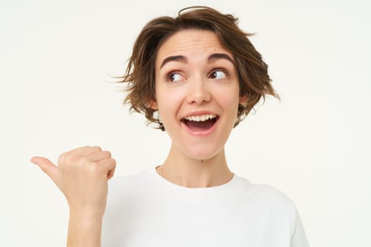 Image of excited brunette woman with short haircut, pointing left, smiling and looking happy, showing you advertisement, standing over white background.