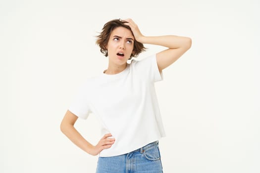 Portrait of complicated woman, touches her head, looking up, puzzled by something, feeling distressed, has problem, stands over white background.