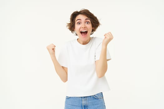 Excited brunette girl, student winning prize, celebrating victory, triumphing, standing in white t-shirt and jeans over white background.