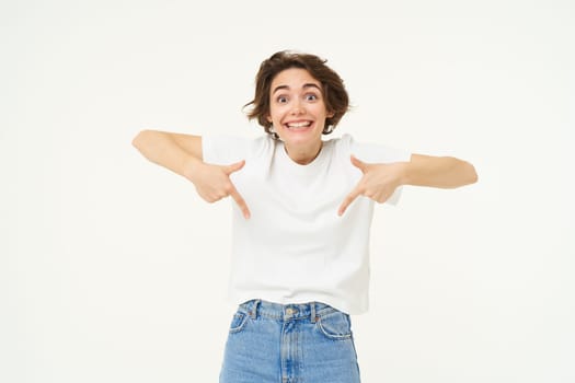 Portrait of cute, happy young woman pointing fingers down, showing advertisement, demonstrating banner on the bottom, standing against white background.