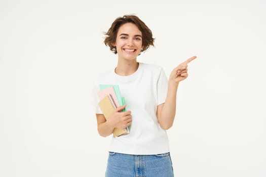 Portrait of brunette woman laughing, student with notebooks pointing at upper right corner, showing banner or advertisement, standing over white studio background.