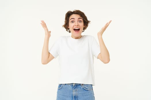 Portrait of amazed, happy young woman, raising hands up and looks surprised, standing excited against white background.