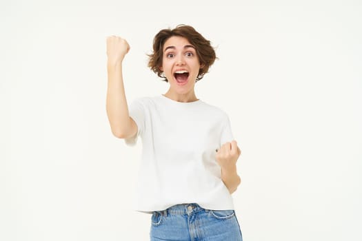 Excited brunette girl, student winning prize, celebrating victory, triumphing, standing in white t-shirt and jeans over white background.